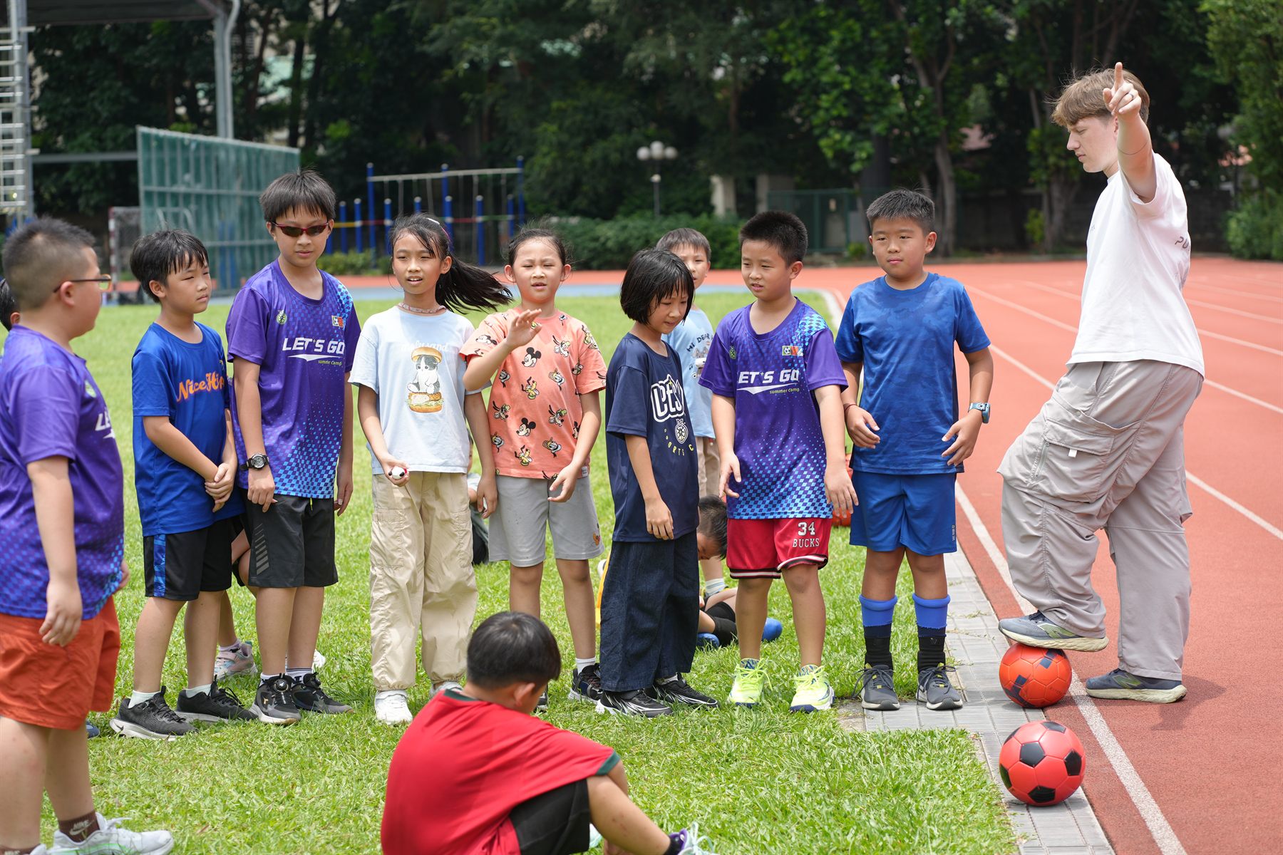 A coach gives instructions to children in Let’s Go summer camp jerseys on a school field