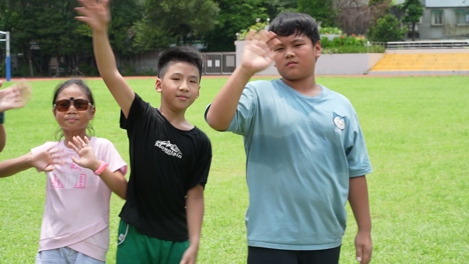 Children on a school field singing together, hands raised, following the motions of a song
